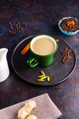 Fototapeta na wymiar Masala chai tea. Traditional indian drink - masala tea with various spices on a black plate. Ceramic cup of spices tea and milk jug