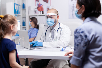 Doctor consulting child patient sitting next to her mother during coronavirus pandemic and wearing face mask. Specialist in medicine providing health care services examination, radiographic treatment