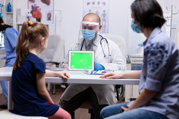 Fototapeta premium Doctor with face mask against coronavirus holding green screen tablet in medical office sitting on the desk. Pediatrician doctor in protection gloves and mask providing clinic medicinal health care