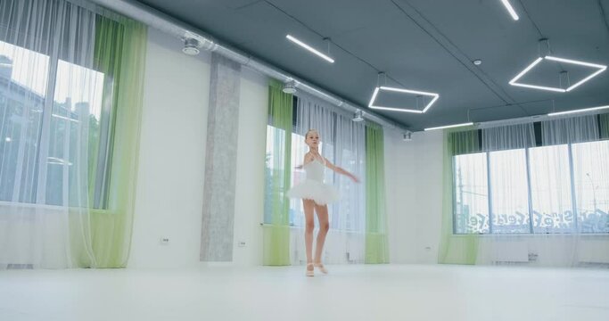 beautiful little ballerina in white suit with tutu performs classical dance in studio with large windows