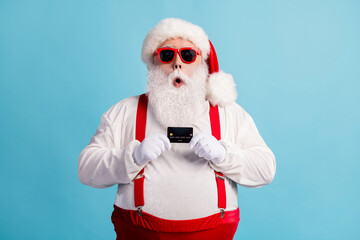 Fototapeta na wymiar Photo of pensioner grandfather grey beard hold debit card suggest advice shopping service wear santa x-mas costume red suspenders sunglass white gloves cap isolated blue color background