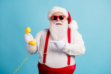 Fototapeta na wymiar Portrait of his he nice attractive cheerful funky white-haired Santa holding in hand demonstrating yellow retro telephone isolated over bright vivid shine vibrant blue color background