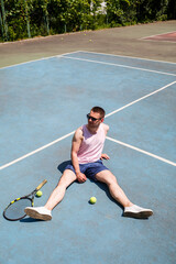 Handsome man in a T-shirt and shorts sits on a tennis field with a racket. Guy in sunglasses