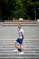Obraz premium A handsome man in a T-shirt and shorts stands on the steps. Guy in sunglasses