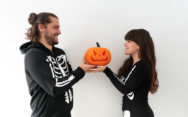 A couple of a boy and a girl dressed as skeletons holding a halloween pumpkin while they look into each other's eyes and smile