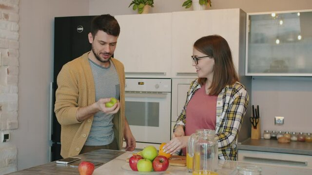 Cheerful young man is juggling with apple while pregnant wife is laughing cooking in kitchen at home. Happy family and leisure activities concept.