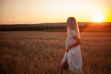 Fototapeta na wymiar A young blonde woman walks around the evening field and looks at the sunset. Village life, slow life