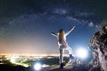Fototapeta na wymiar Back view of space traveler spreading arms out to sides while standing on rocky hill under beautiful sky with stars. Mission specialist astronaut in space suit enjoying view of milky way, night city
