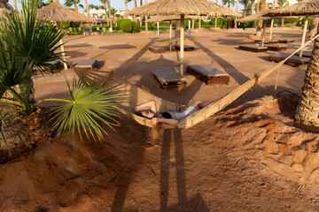 Happy girl relaxing on beach hammock in tropical vacation resort hotel. Holidays in the tanning under the sun. Woman wearing sunglasses relax lying down on sun lounger bed.