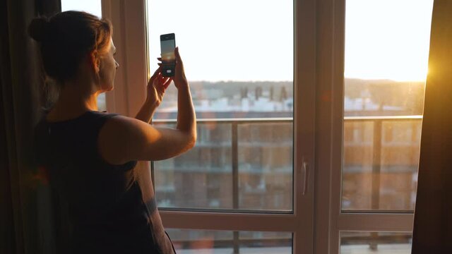 Woman stands near the window and takes pictures of the setting sun on a smartphone