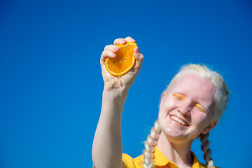 An albino young woman in a yellow blouse against a blue sky. holding slices of sliced orange.