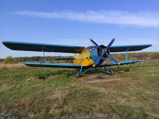 Yellow and blue old biplane plane with a single piston engine and propeller against a blue sky with clouds on the airfield with green grass in summer