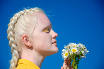 A young albino woman in a yellow blouse against a blue sky. holding a bouquet of daisies in her...