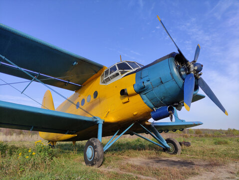 Yellow and blue old biplane plane with a single piston engine and propeller against a blue sky with clouds on the airfield with green grass in summer