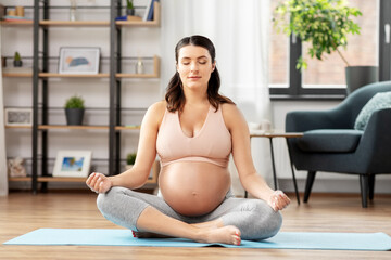 sport, yoga and people concept - happy pregnant woman with earphones and smartphone listening to music and meditating at home