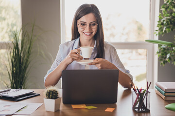 Photo of charming lady sit desktop arms hold cup saucer beaming smile look screen pc wear specs...