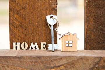 Keychain of wooden figure house on fence with key and inscription in English letters "home". Cottage in the background. Building, project, moving to new house, mortgage, rent and purchase real estate.