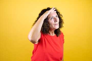 Middle age woman wearing casual shirt standing over isolated yellow background very happy and smiling looking far away with hand over head. Searching concept.