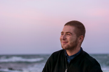 Smiling young Caucasian man with beard on background of the sea and waves with white foam. Selective focus, close-up