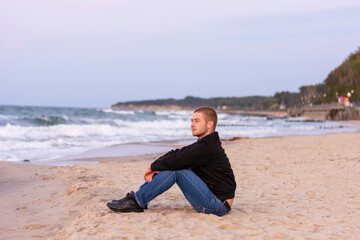 Fototapeta na wymiar Young handsome man with a beard is resting on seashore in evening at sunset. Romantic mood, selective focus