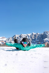 A teenage girl in a colorful ski suit, helmet and sunglasses lies in the snow and smiles happily against the backdrop of mountain peaks. Sports concept, people, travel.