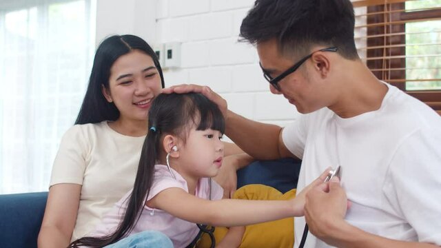 Happy cheerful Asian family dad, mom and daughter playing funny game as doctor having fun on sofa at home. Self-isolation, stay at home, social distancing, quarantine for coronavirus prevention.