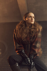 Obraz na płótnie Canvas stylish curly girl in a vintage checkered jacket, leather gloves and black pants sits on a chair in the loft studio and looks away
