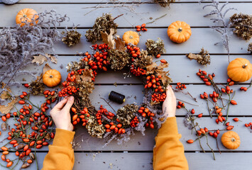 Girl making floral autumn door wreath using colorful rosehip berries, rowan, dry flowers and pumpkins. Fall flower decoration workshop, florist at work.
