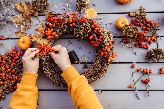 Girl making floral autumn door wreath using colorful rosehip berries, rowan, dry flowers and pumpkins. Fall flower decoration workshop, florist at work.