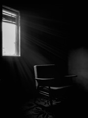Dramatic room in abandoned house with an old empty chair near the window, black and white image. Light coming from the window, vertical shot.