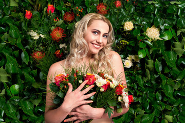 A beautiful young blonde lady standing in front of a wall of flowers holding a bouquet
