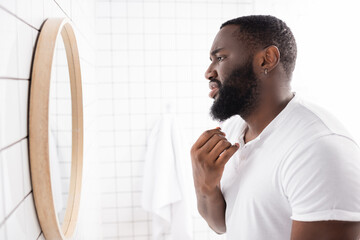 side view of unhappy afro-american man looking at beard in mirror