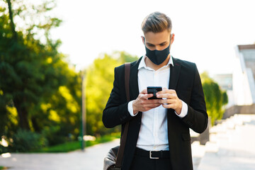 Handsome businessman in face mask using mobile phone on city street