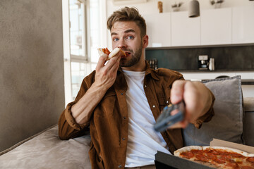 Excited guy eating pizza while watching tv and using remote control