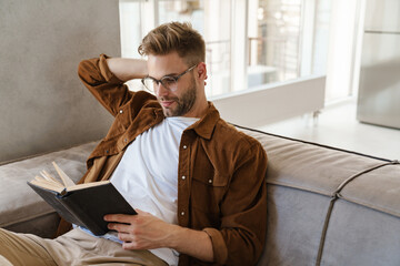 Handsome focused guy in eyeglasses reading book while sitting on sofa