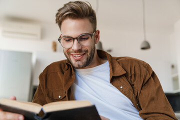 Handsome smiling guy in eyeglasses reading book while sitting on sofa
