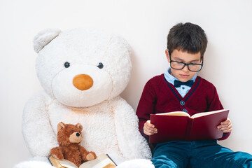 A schoolboy in a sweater and glasses sits next to a large teddy bear and reads a book. Conceptual. Copy space.