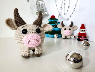 Beige knitted toy decoration of a bull cow in the foreground. For new year, christmas 2021.on a white background with a Christmas tree and santa claus.