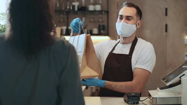 An attractive young guy barista sells coffee to a client behind the counter during quarantine indoors