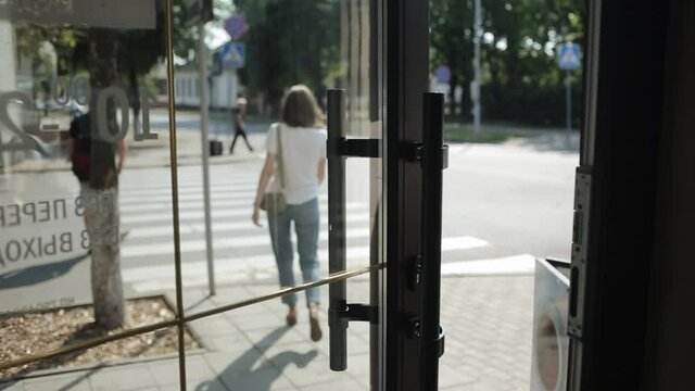 A young woman in a white T-shirt and jeans walks out of the building through a glass door and walks across the road. View from the back. Shooting from inside the building