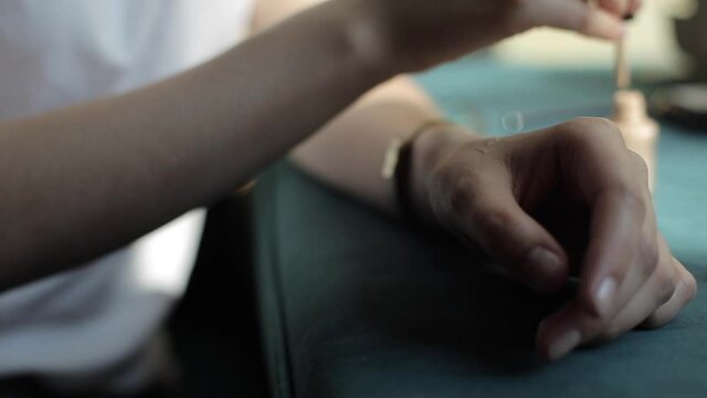 A young woman in a white T-shirt sits at a table and applies foundation from a jar to her hand. Close-up of hands. Blurred background