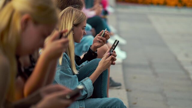 Group of teenagers sitting in row on stairs in city center and using smartphones, focus on blonde girl wearing denim outfit listening to voice message on gadget. Young generation communication