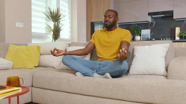 African american calm young man starts to meditate in his living room. Peaceful handsome black guy meditating for concentration, calmness and positive mind.