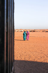 Two Saharawi women walking in the desert together with the refugee camp in the background