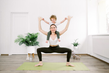 Obraz premium Beautiful young Caucasian woman and her little son are smiling while doing yoga together at home. Woman in yoga position with her son sitting on her shoulders