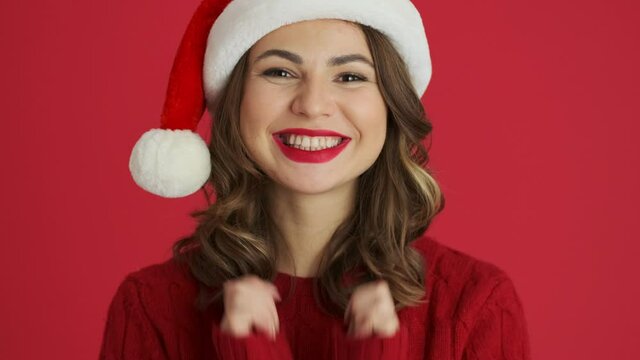 A close-up view of a surprised woman wearing a warm red sweater and christmas hat is looking to the camera while opening her mouth standing isolated over red background