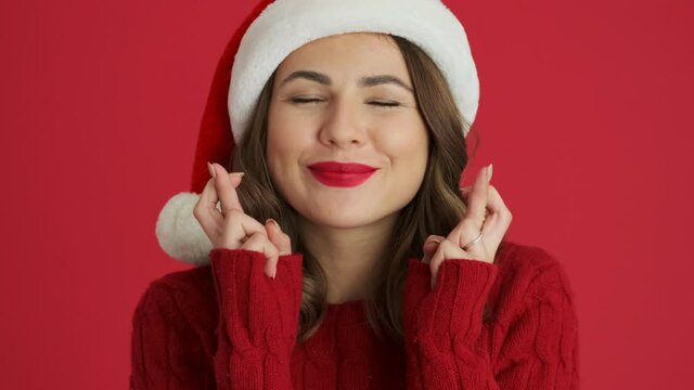A close-up view of a smiling positive woman wearing a warm red sweater and christmas hat is making a wish while crossing her fingers standing isolated over red background