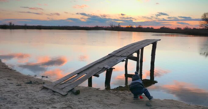 Child Throws Sand in Water near Wooden Fishing Pier on River Lake Pond Shore. Scenic Picturesque Sunset Cloudscape Reflection Beautiful Evening Countryside Landscape. Rural Scene