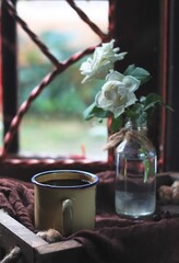 Selective focus of a cup of coffee near the window, looking outside rainy day,  with white roses at clear bottle. Vintage and romantic look. Dark moody photo.  Coffee concept.