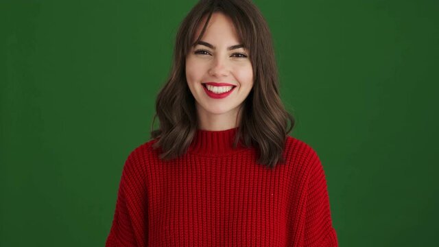 A smiling woman in a red sweater is waving her hand doing hello gesture standing isolated over green background